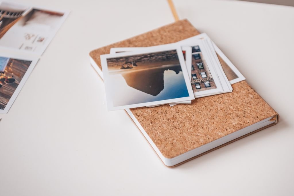 Cork-covered notebook topped with printed photographs on a white desk, conveying travel memories.